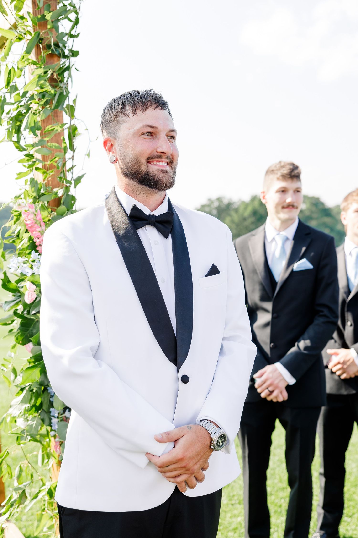 Smiling groom in white tuxedo stands at altar awaiting bride at outdoor wedding.