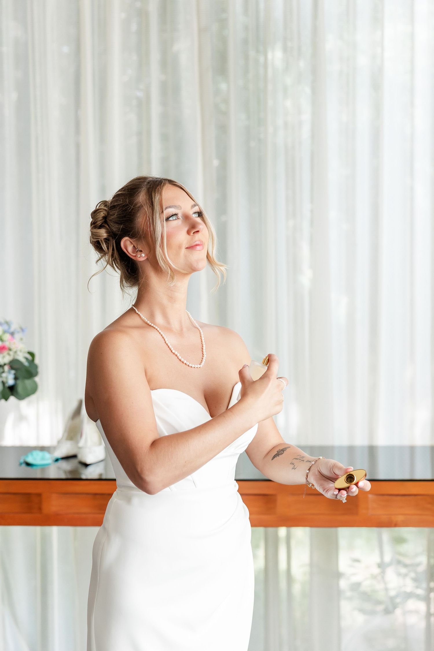 Bride in strapless white gown applying perfume wearing pearl necklace before wedding ceremony.