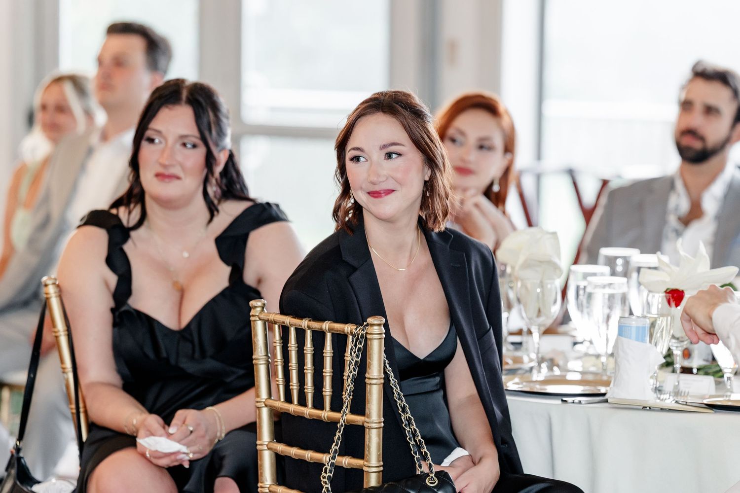 Guests in black attire smile and watch during a wedding reception speech at an elegant venue.