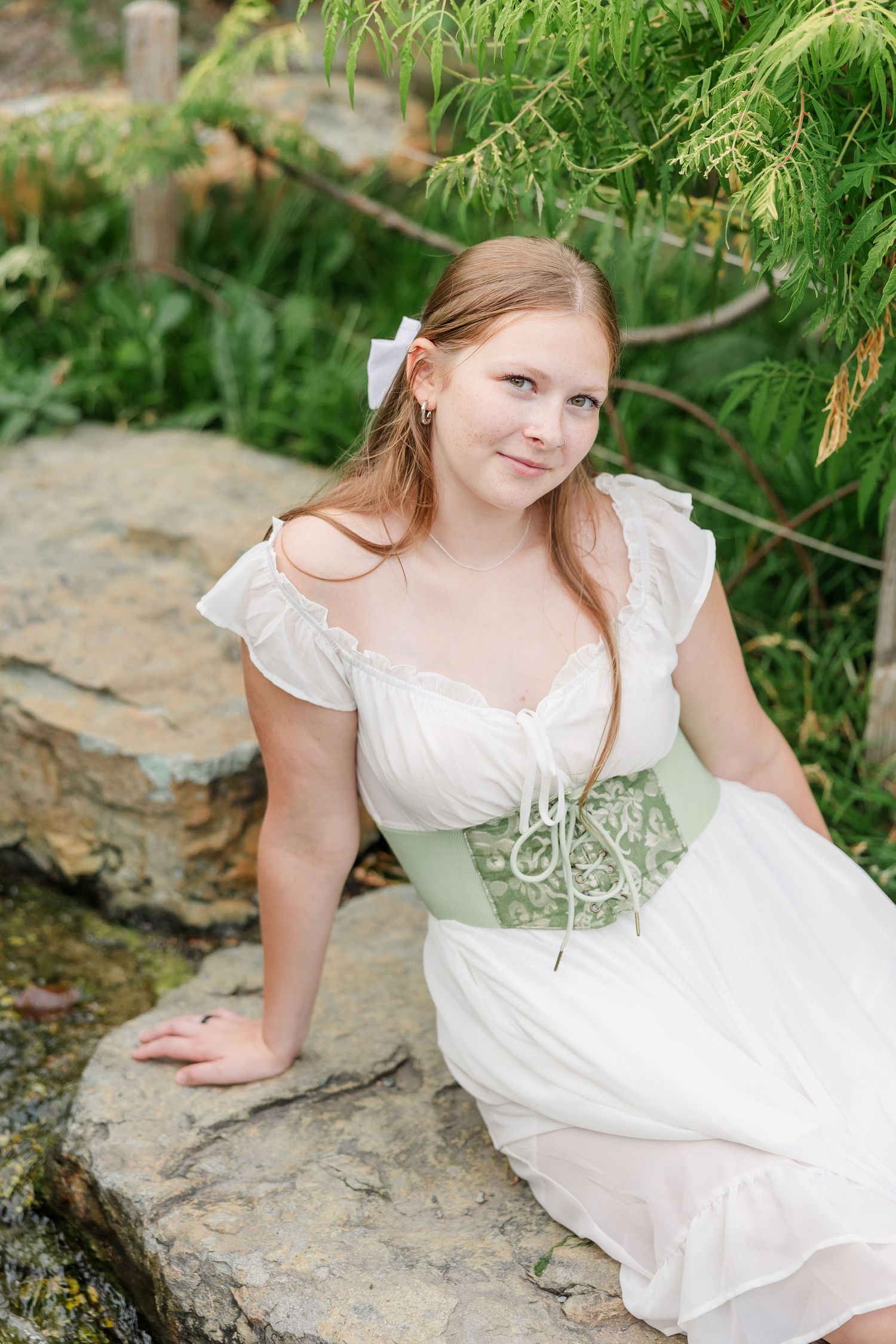 Samantha sits on a garden rock in a white dress and green corset during her whimsical senior session.