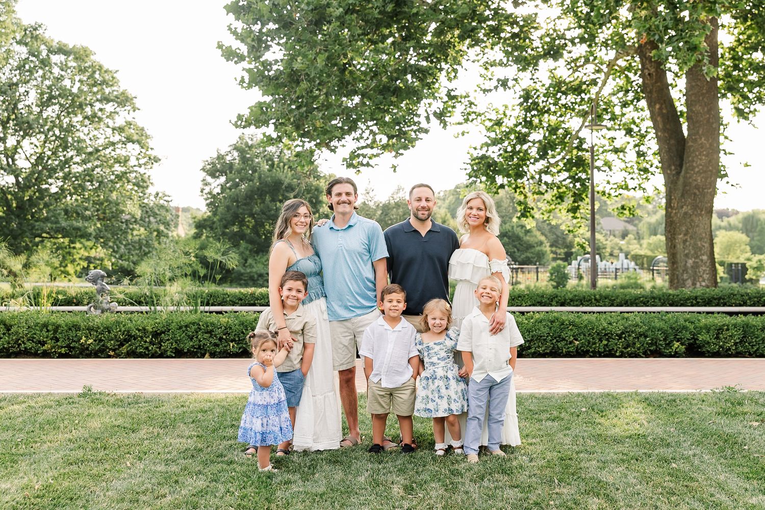 A joyful family of eight poses outdoors on green grass with manicured hedges and trees in the background.