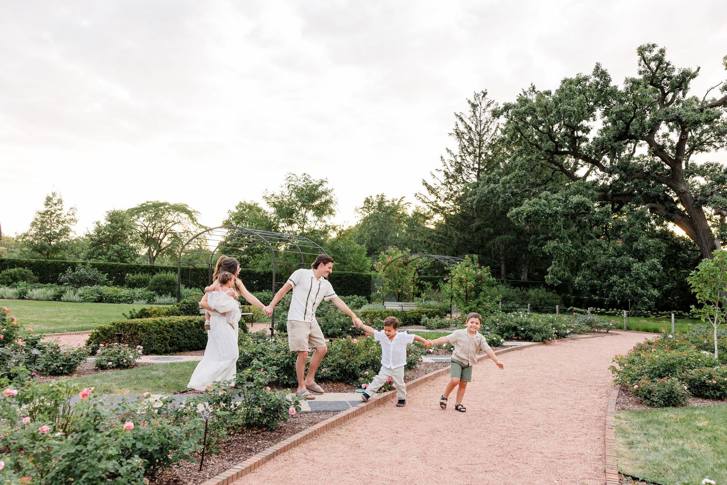 A family of four holds hands and walks together along a brick garden path surrounded by lush greenery.