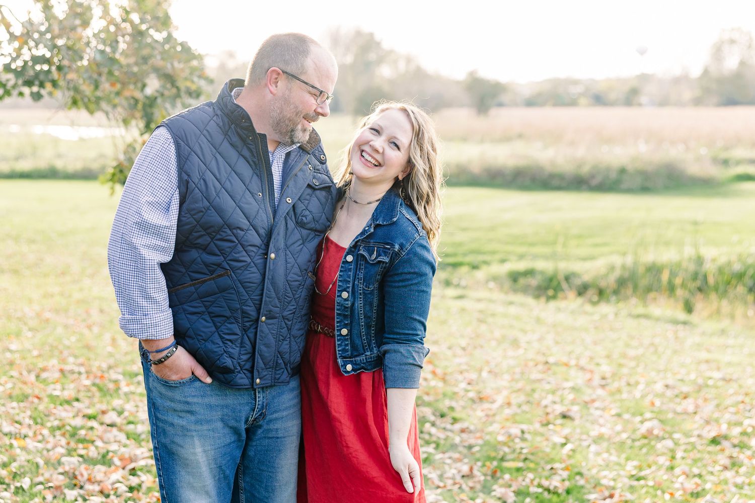 Laughing couple in a vest, denim jacket and red dress standing close together in an open field at golden hour.