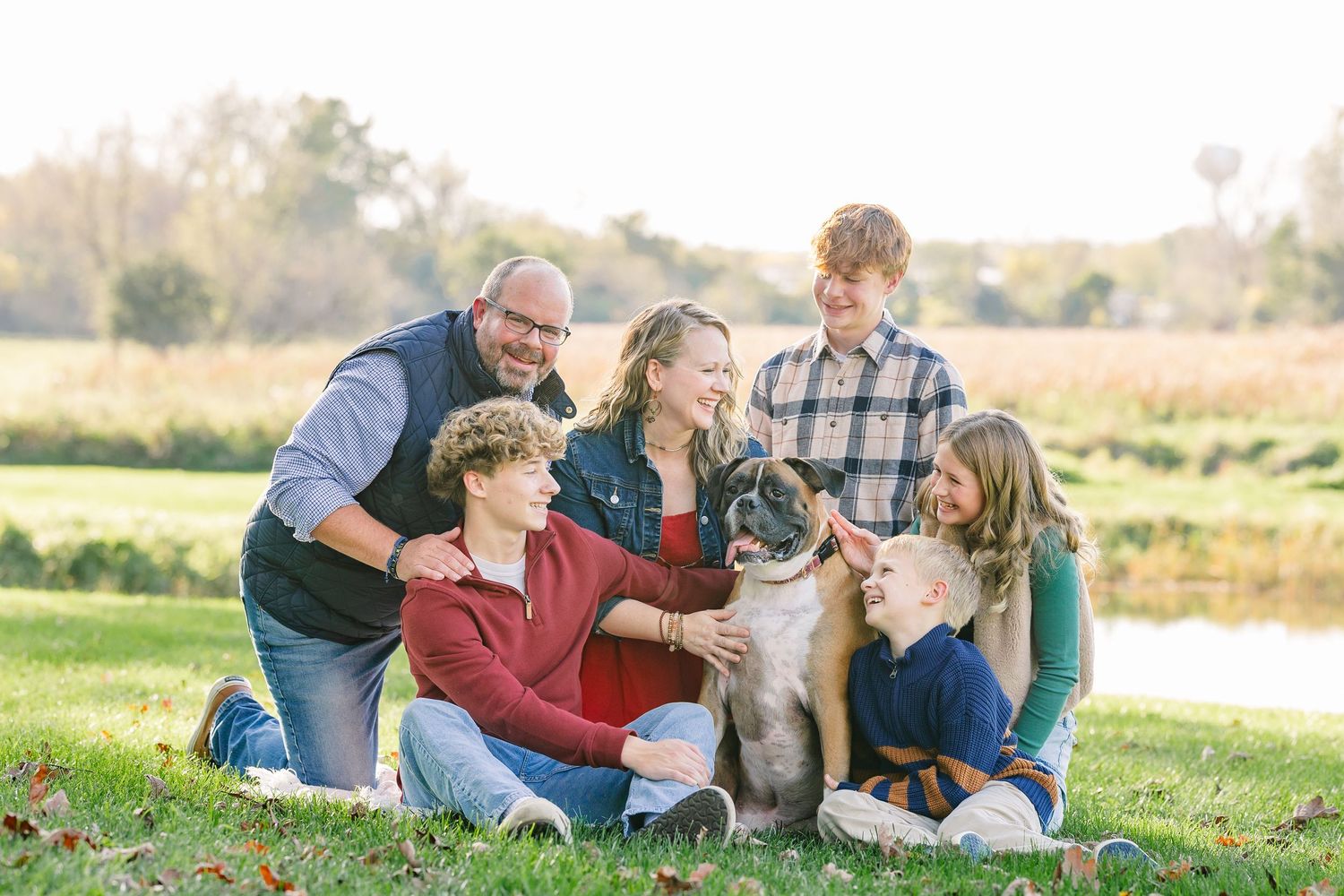 A family of six, including a dog, sits and stands together on a grassy lawn near a pond in warm golden light for an outdoor portrait.