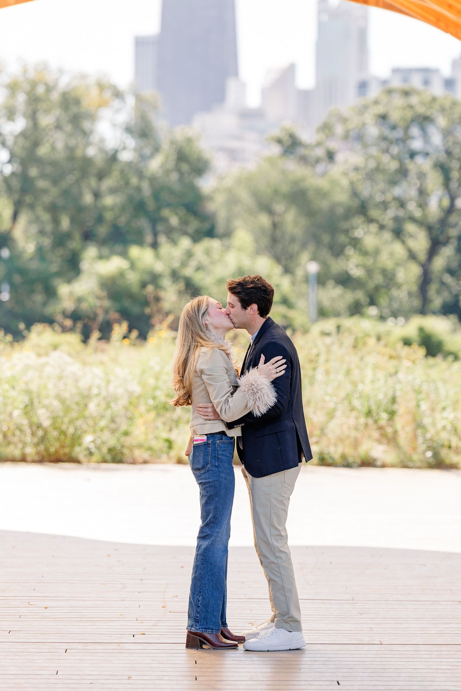 Couple sharing a romantic kiss on a wooden deck with Chicago skyline and lush greenery in the background.