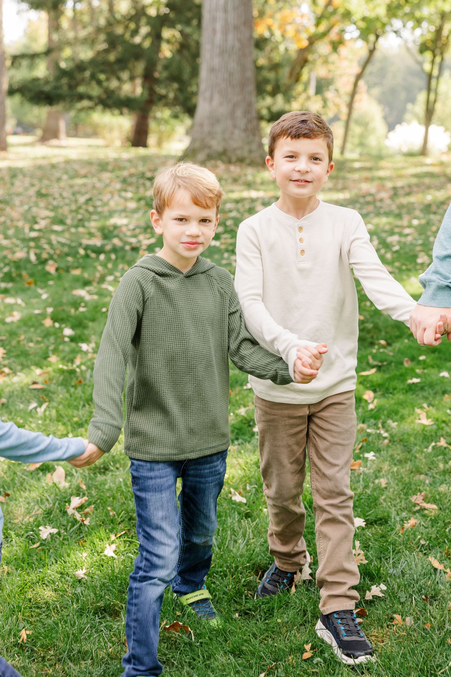 Two young boys holding hands and smiling while standing on grass covered with fallen leaves.