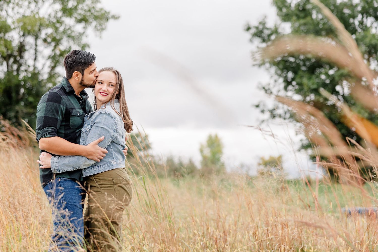 Smiling couple embracing in a tall golden grass field, the man kissing her cheek, with trees and an overcast sky behind.