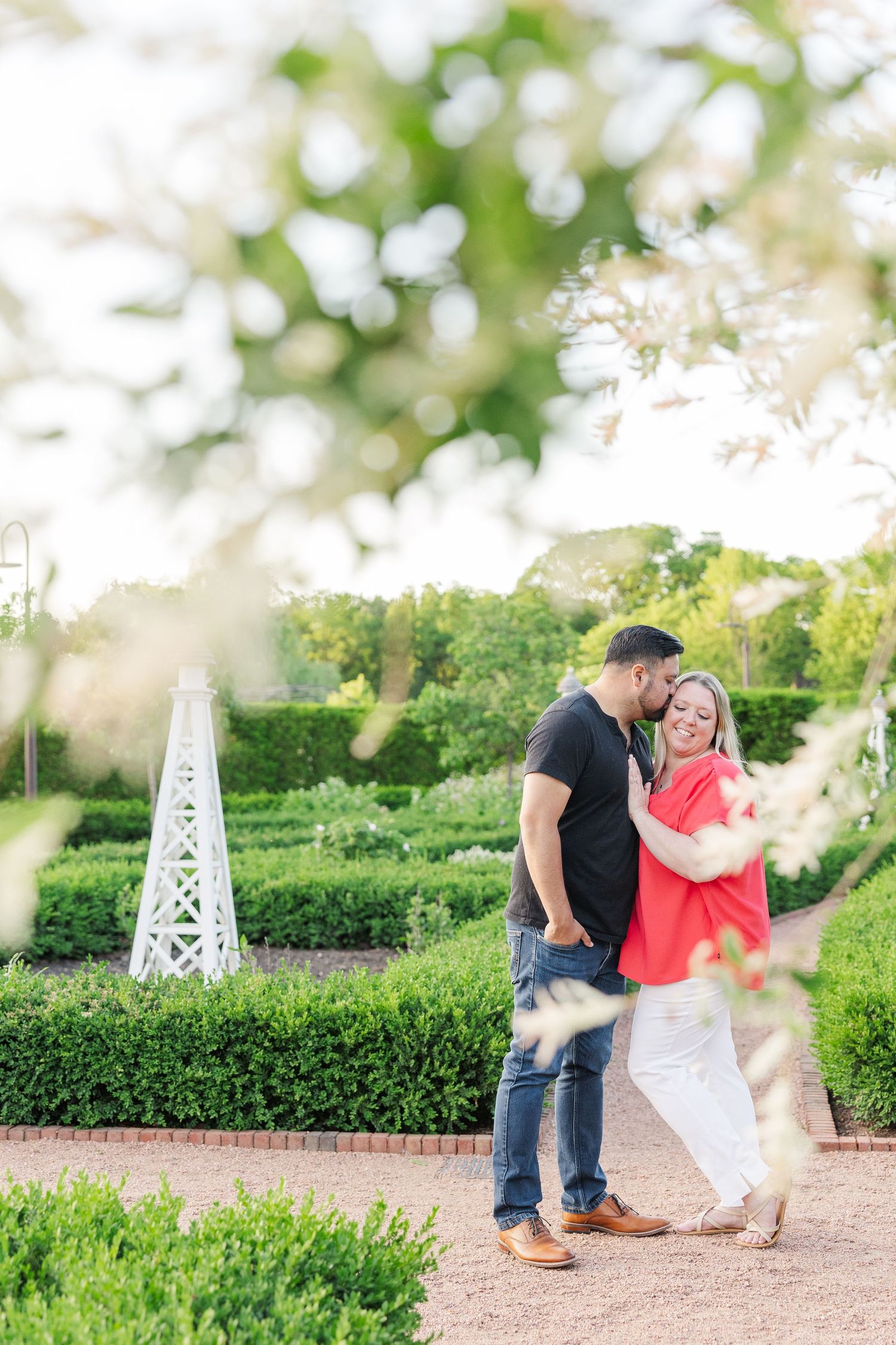 Romantic couple posing in front of a decorative white garden obelisk and landscaped shrubs.