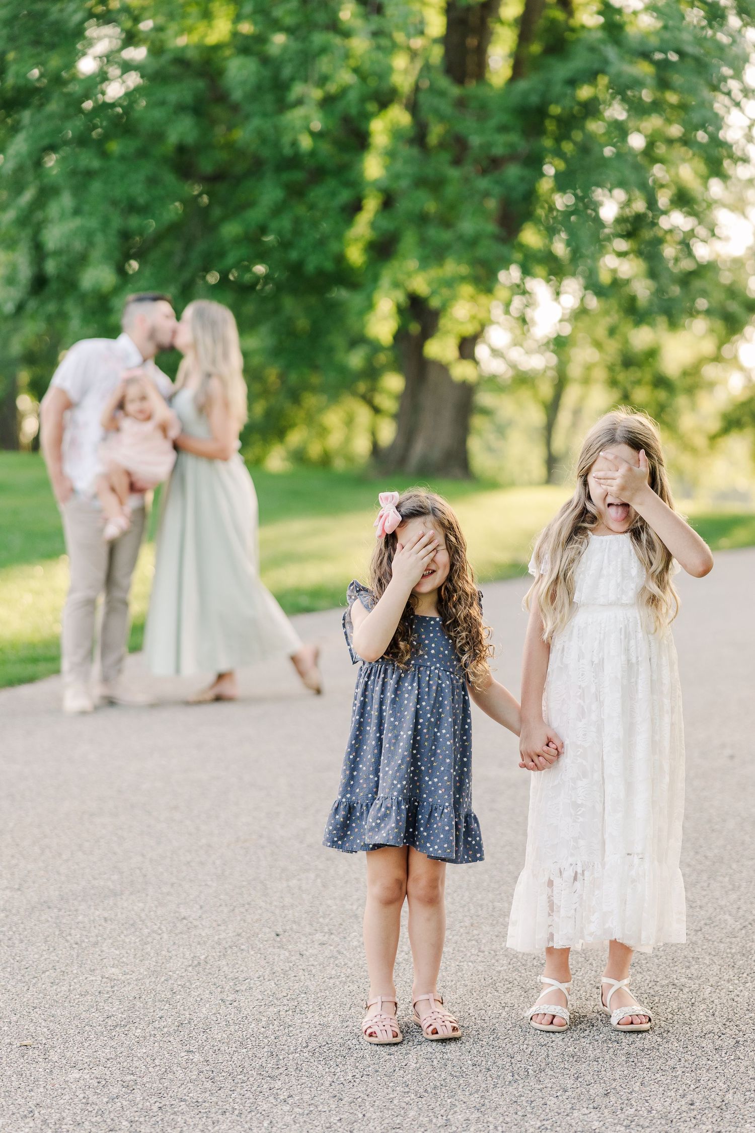 Two little girls playing peek-a-boo on a park path with blurred figures in background.