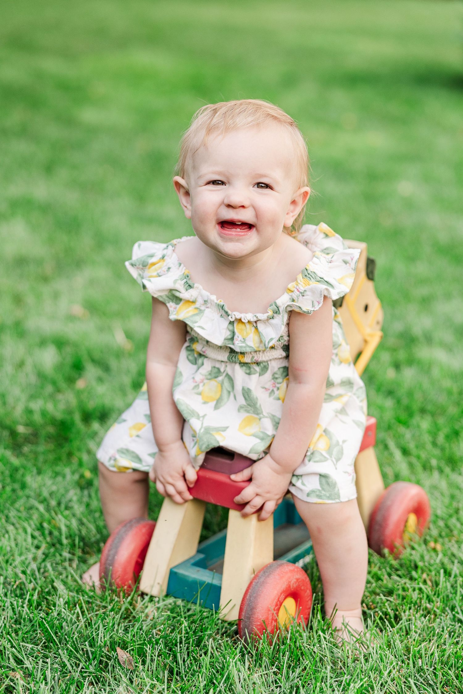 A happy child in a lemon print dress sits on a vintage wooden riding toy in a sunny garden.