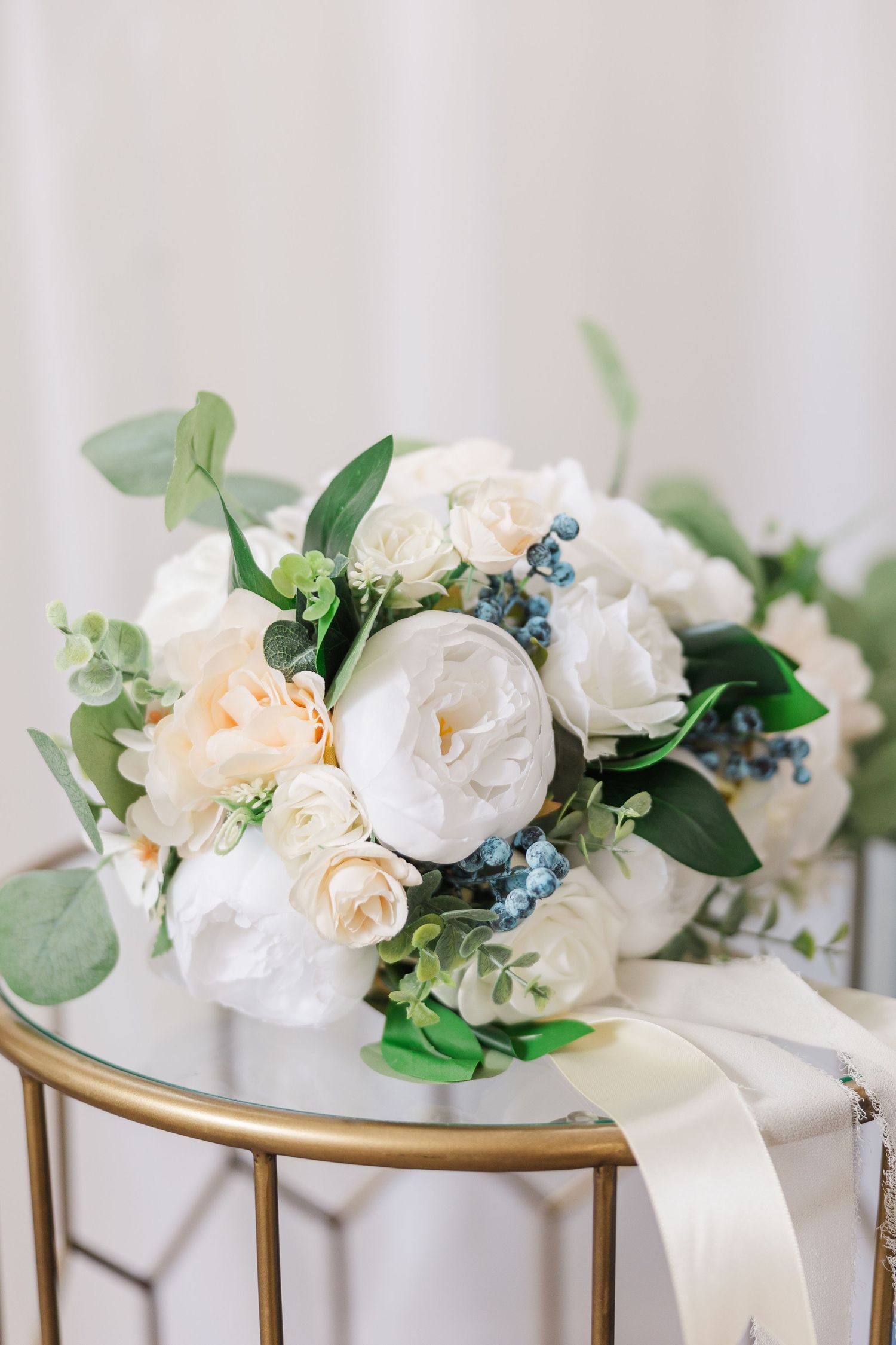Elegant white and peach bridal bouquet with roses, peonies and blue berries on a gold accent table.