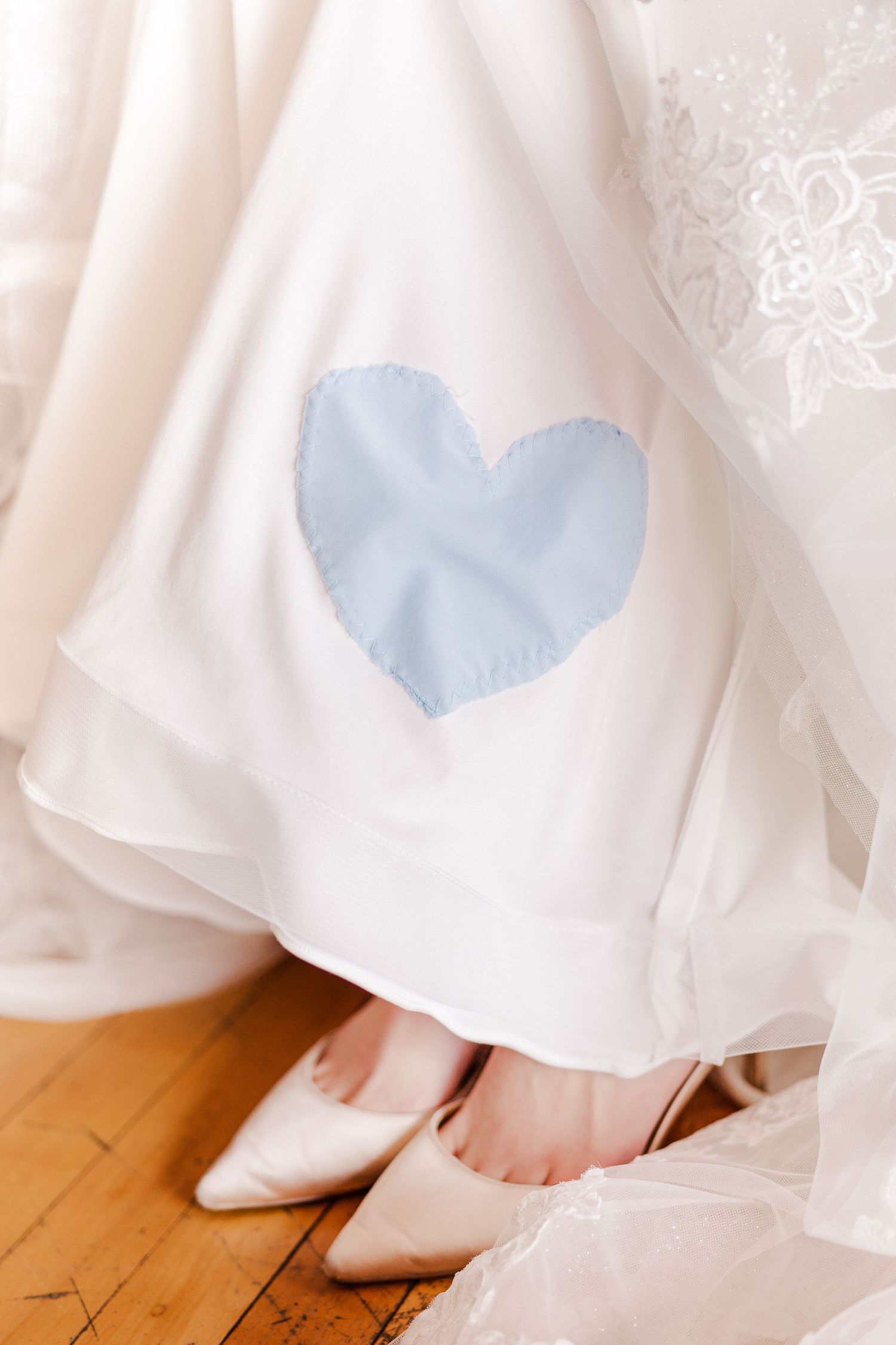 A white wedding dress with a light blue heart detail peeks out above cream colored heels on a hardwood floor.