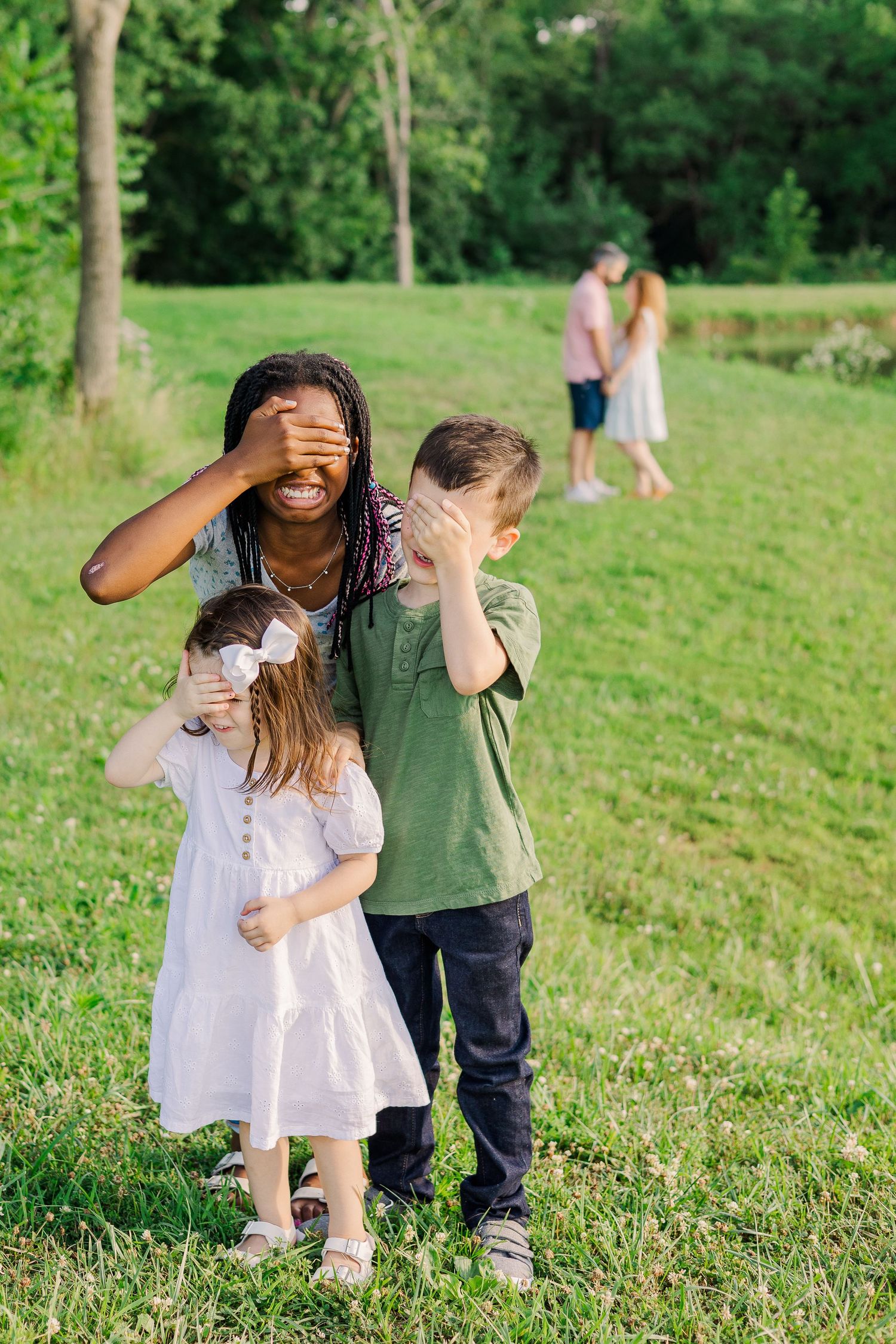 Three people playing hide and seek in a green park with others visible in the background.