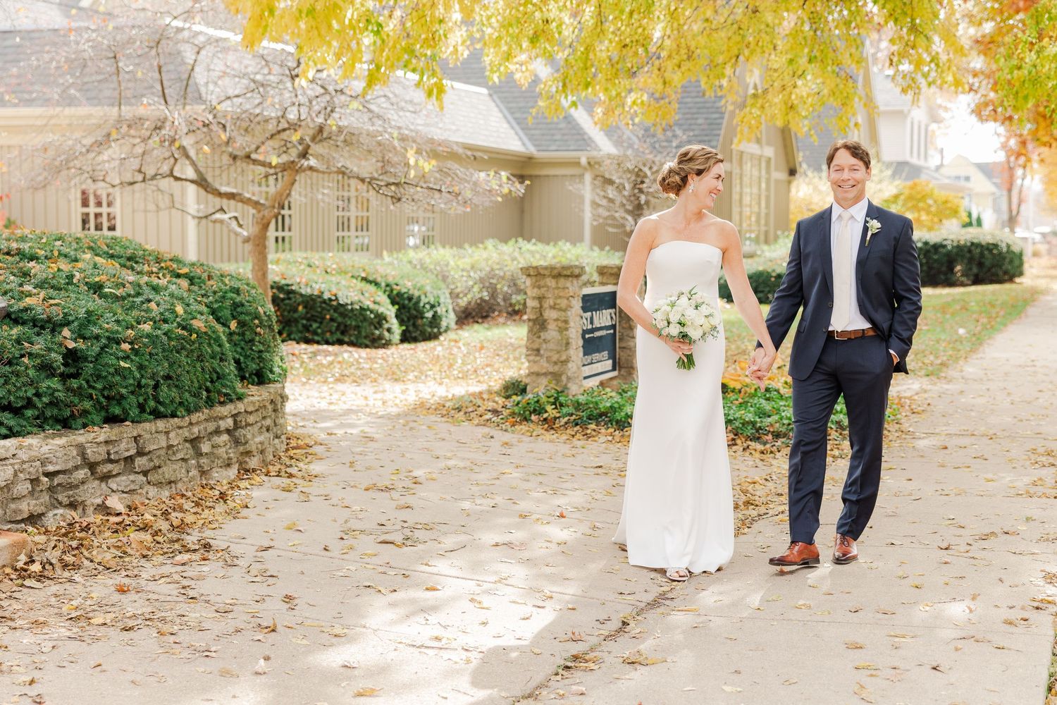 A sunny autumn wedding photo of a couple walking along a stone-lined garden path.