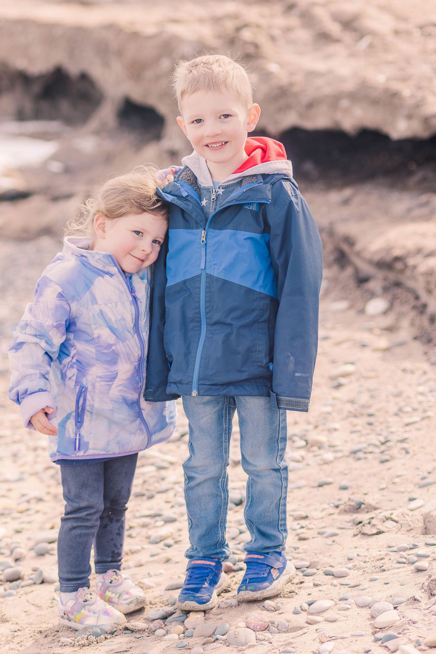 Two children in winter jackets standing together on rocky beach.