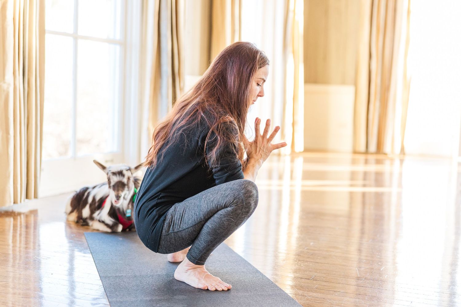 A yoga practitioner performs a deep squat position on a black mat while a dog observes nearby.