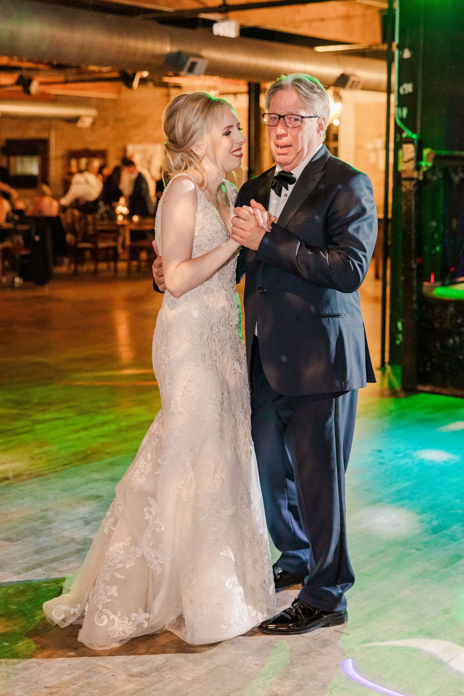 A tender dance between a bride in a lace gown and partner in a navy suit at a dimly lit reception venue.