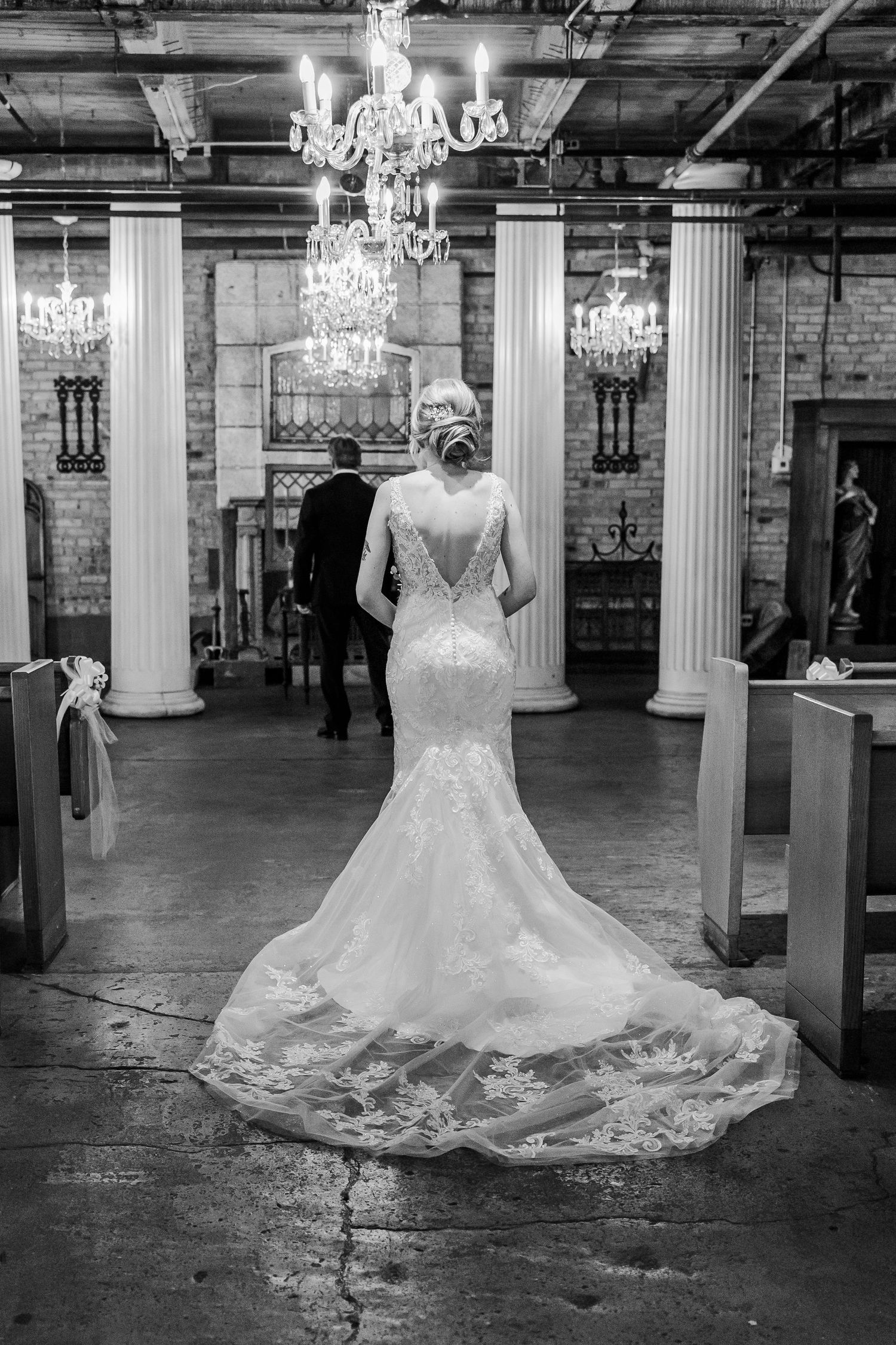 A black and white photo showing an elegant wedding dress with a long train in a chandelier-lit venue.