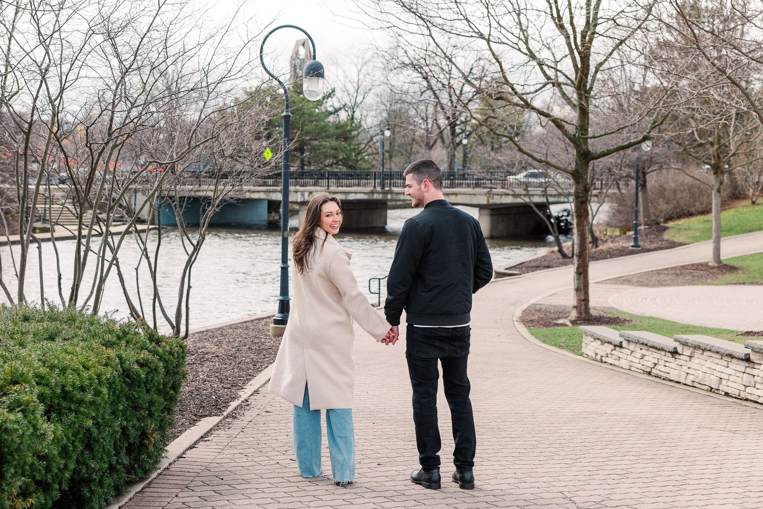 A couple walking together on a paved path near a bridge on an overcast day.