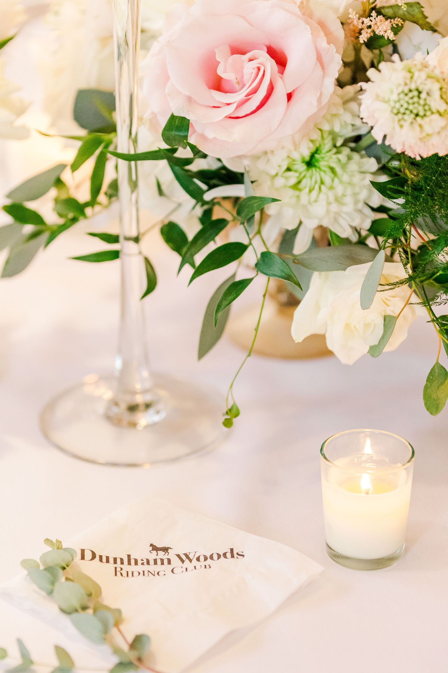 Elegant table setting with pink roses, white flowers, eucalyptus leaves, and a glowing votive candle on a white tablecloth.