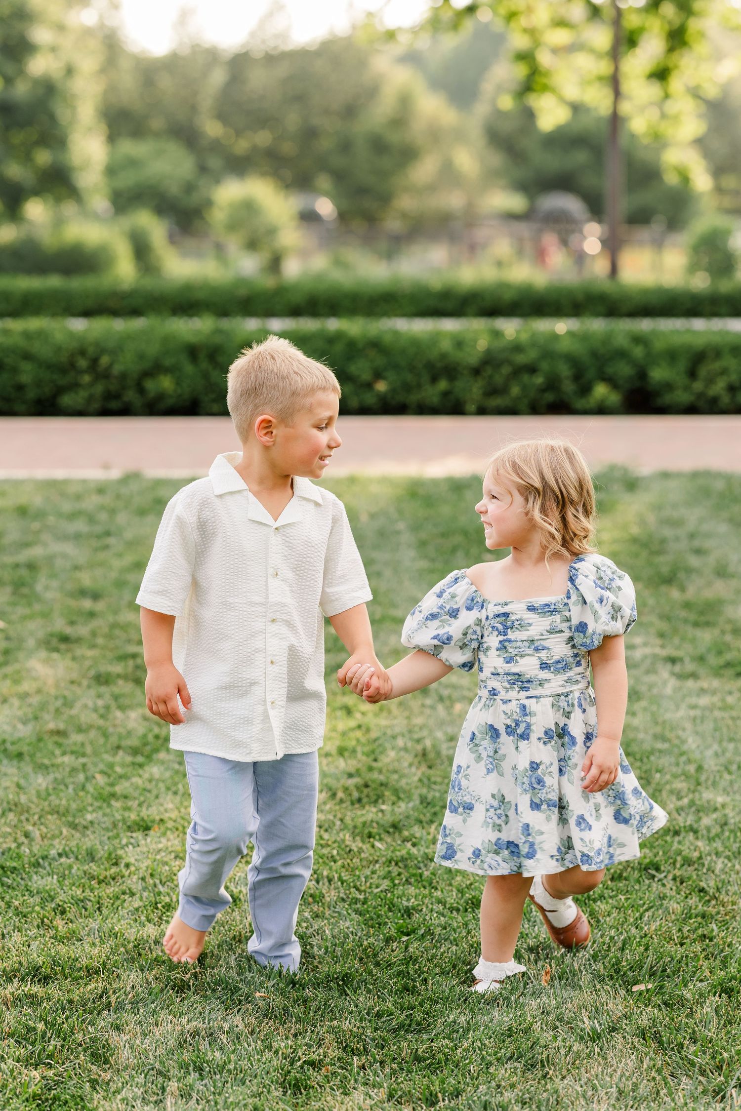 A young boy in a white shirt and a little girl in a blue floral dress hold hands while walking on green grass.