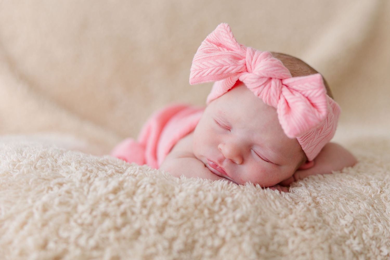 Peaceful newborn baby girl sleeping on fluffy cream blanket wearing pink swaddle and matching bow headband.