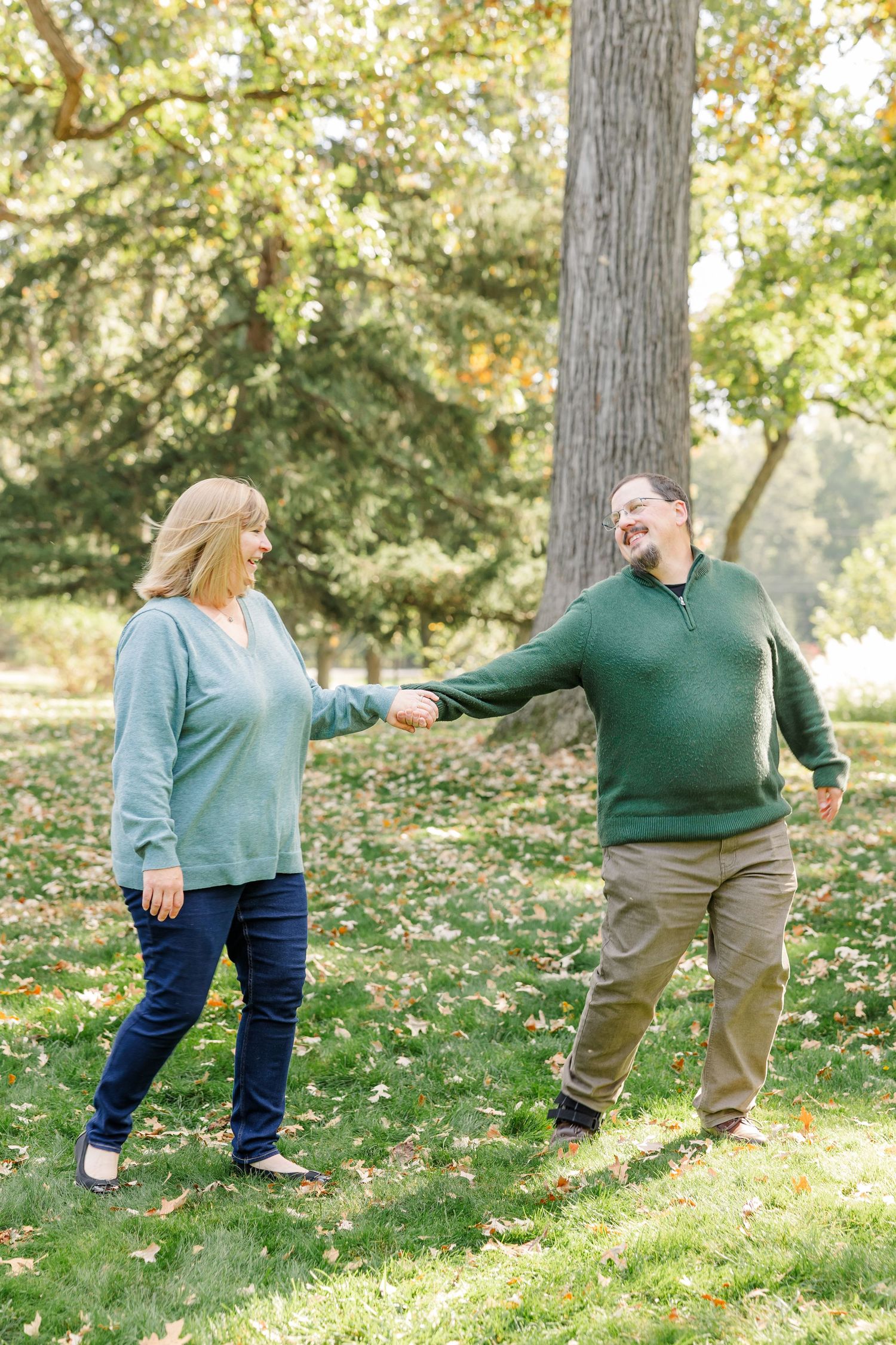 A couple holds hands and smiles while walking together in a lush green park during autumn.