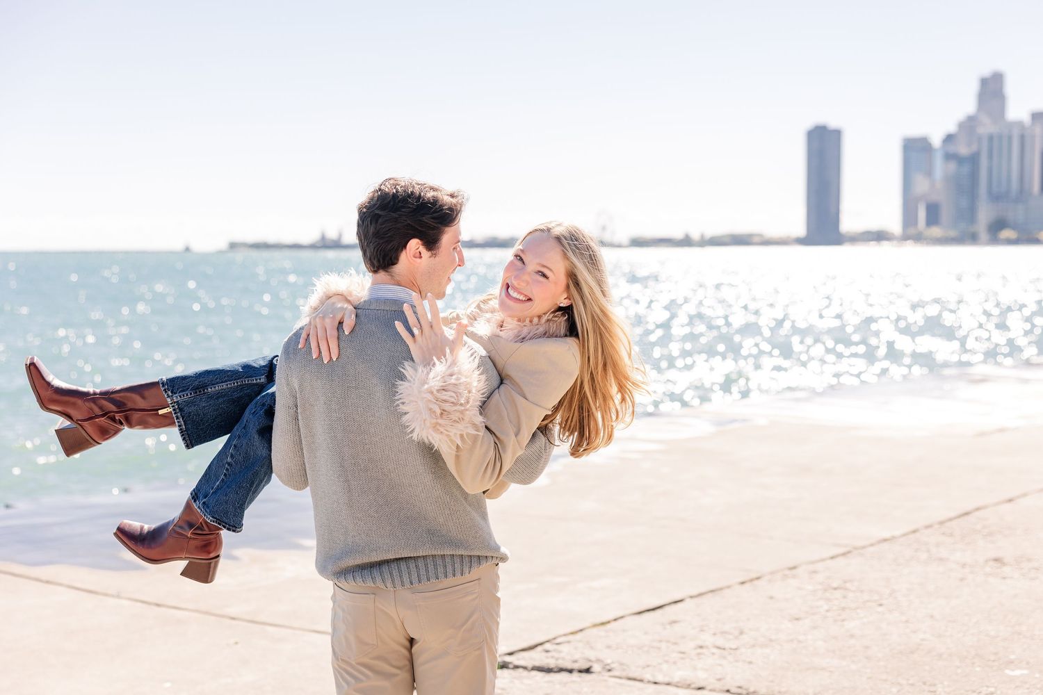 A joyful couple embraces on a lakeside promenade with a city skyline in the background on a sunny day.