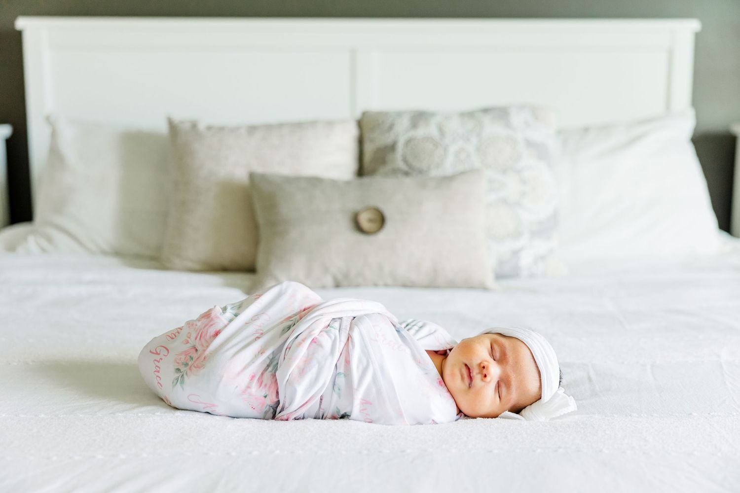 Swaddled newborn baby sleeping peacefully on a white bed with decorative pillows in a bright nursery.