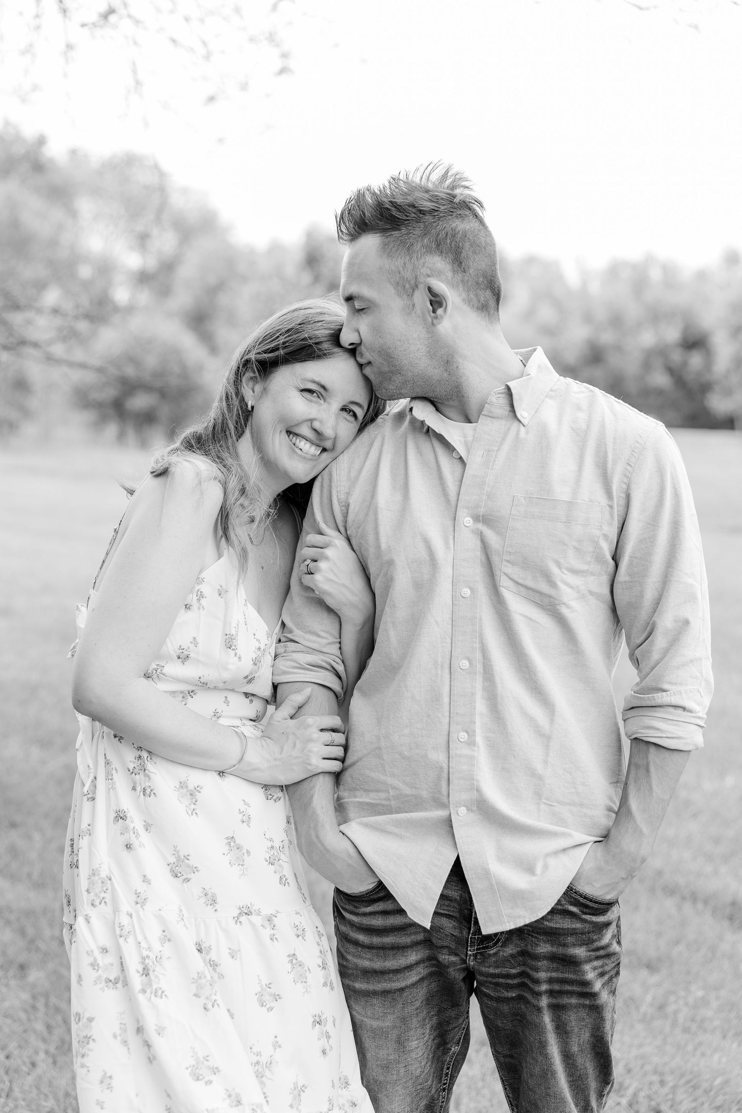 A black and white photo of a man kissing a smiling woman on the head in an outdoor setting.