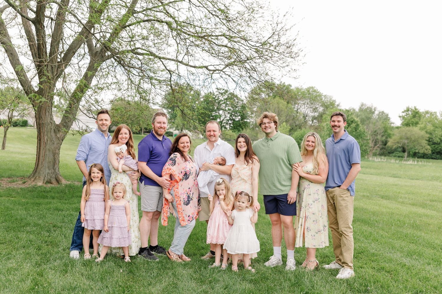 Large family group portrait outdoors on green lawn with trees, wearing pastel spring outfits, smiling together.