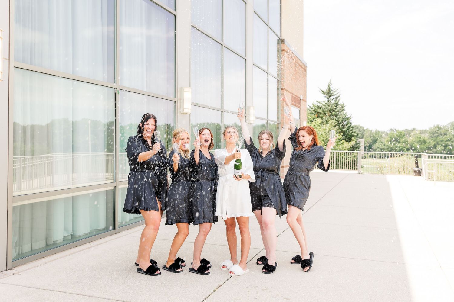 Bride in white dress celebrates with bridesmaids in black robes popping champagne outside modern venue.