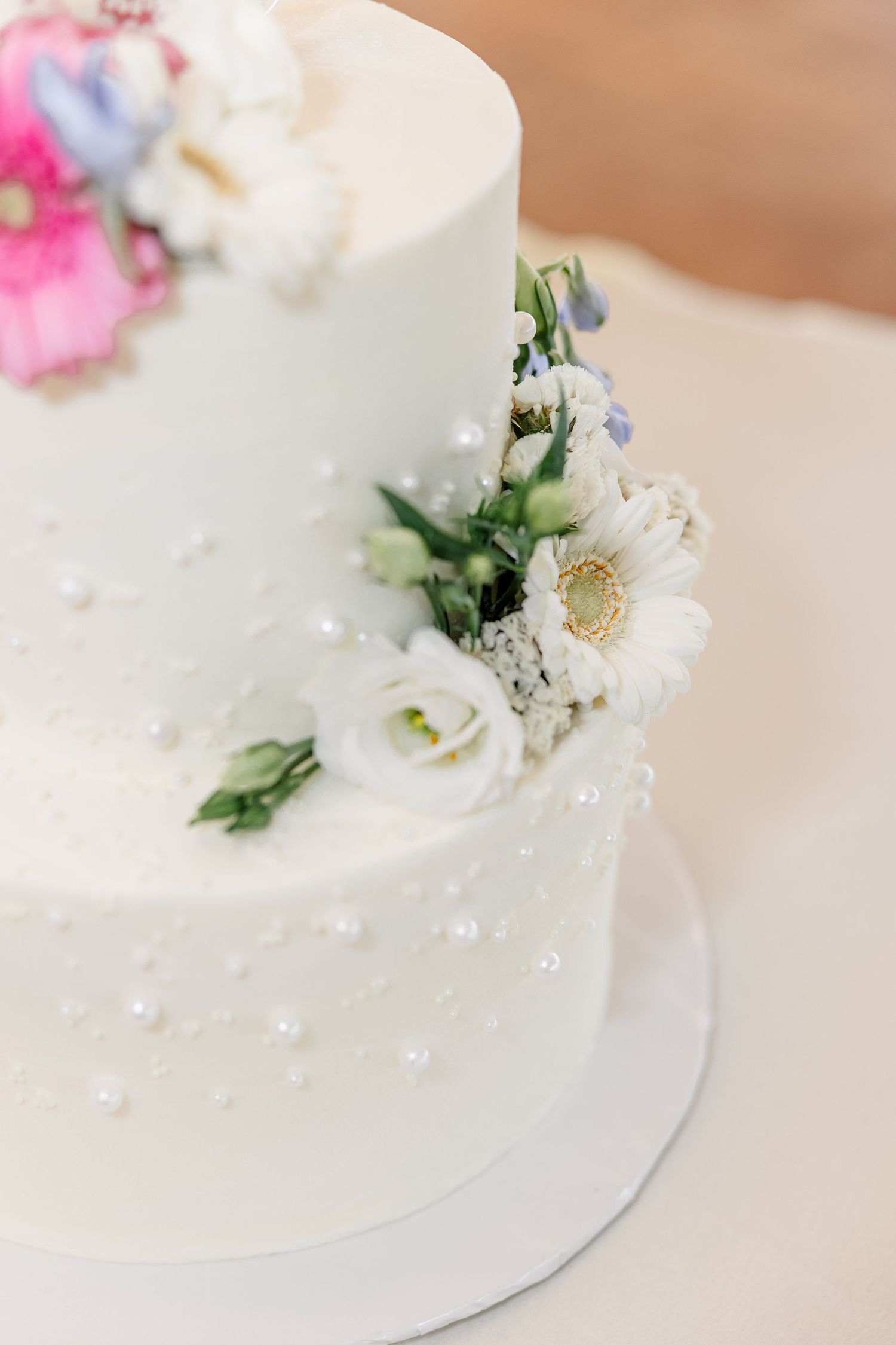Elegant two-tier white wedding cake decorated with fresh white flowers, daisies, and pearl details.