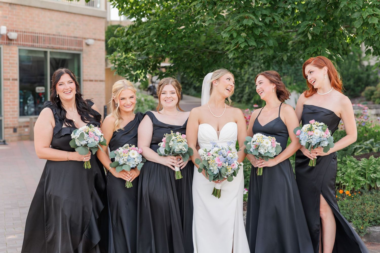 Bride with five bridesmaids in black gowns holding bouquets outdoors on wedding day.