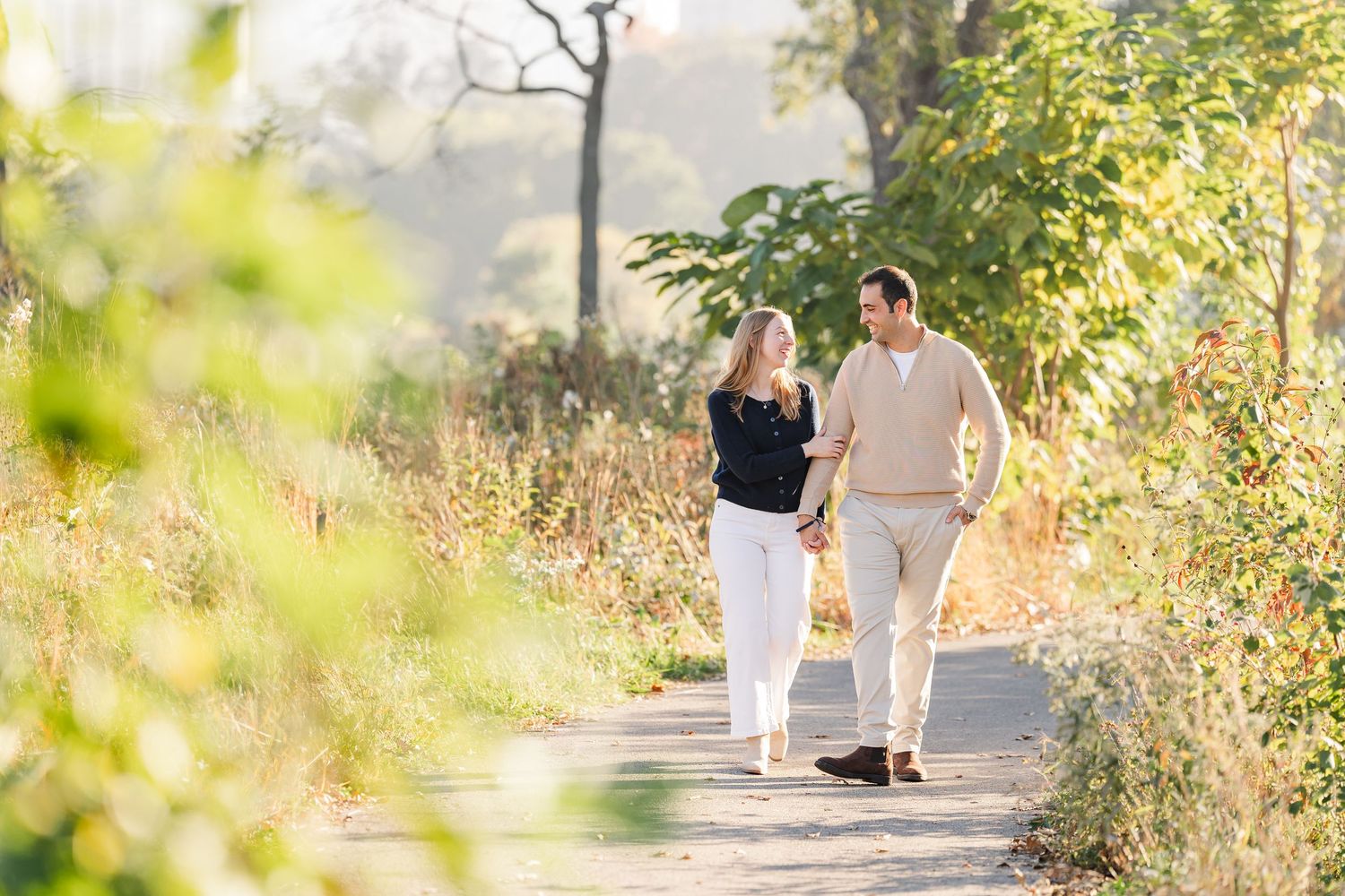Couple holding hands and walking along a sunlit dirt path surrounded by tall golden grasses and trees during an outdoor engagement photo ses