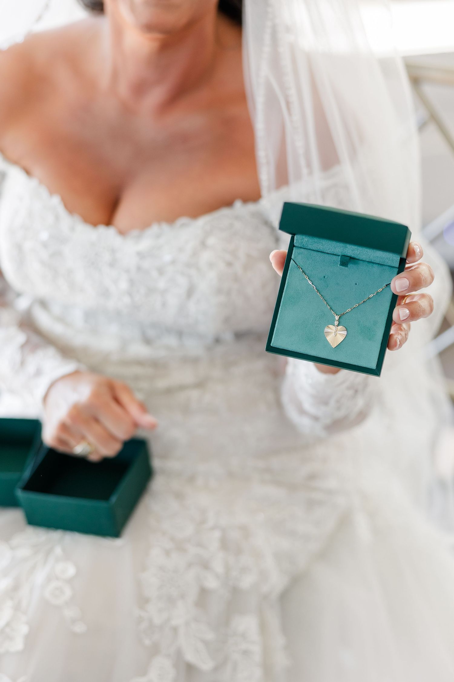 Close-up of a bride in a lace wedding dress holding an open green gift box with a silver heart pendant necklace inside.