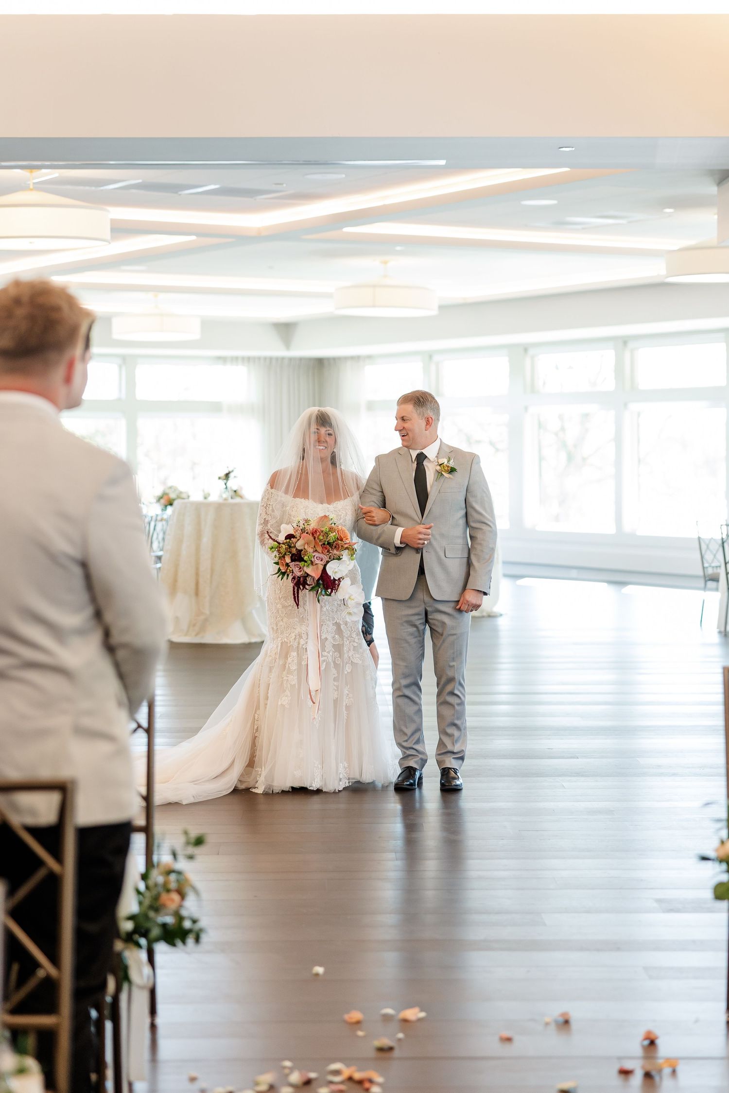 A bride in a lace gown and veil holding a bouquet walks down the aisle on a man's arm at an indoor wedding ceremony.