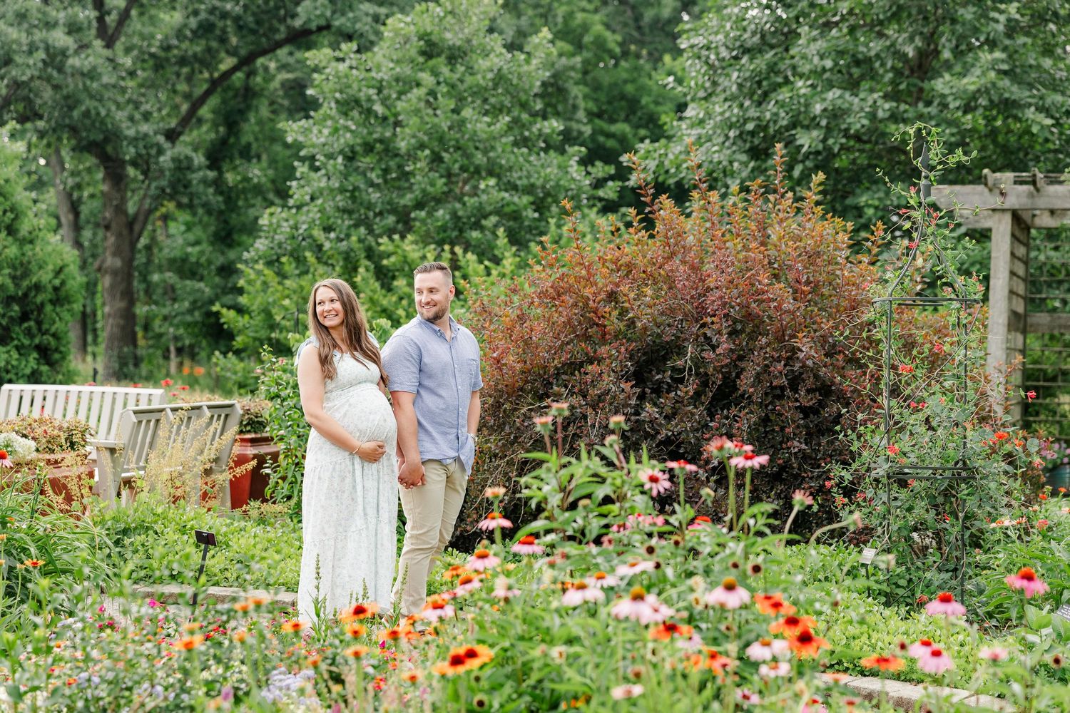 Pregnant couple stands hand-in-hand in colorful flower garden with wooden pergola and vibrant blooms surrounding them.