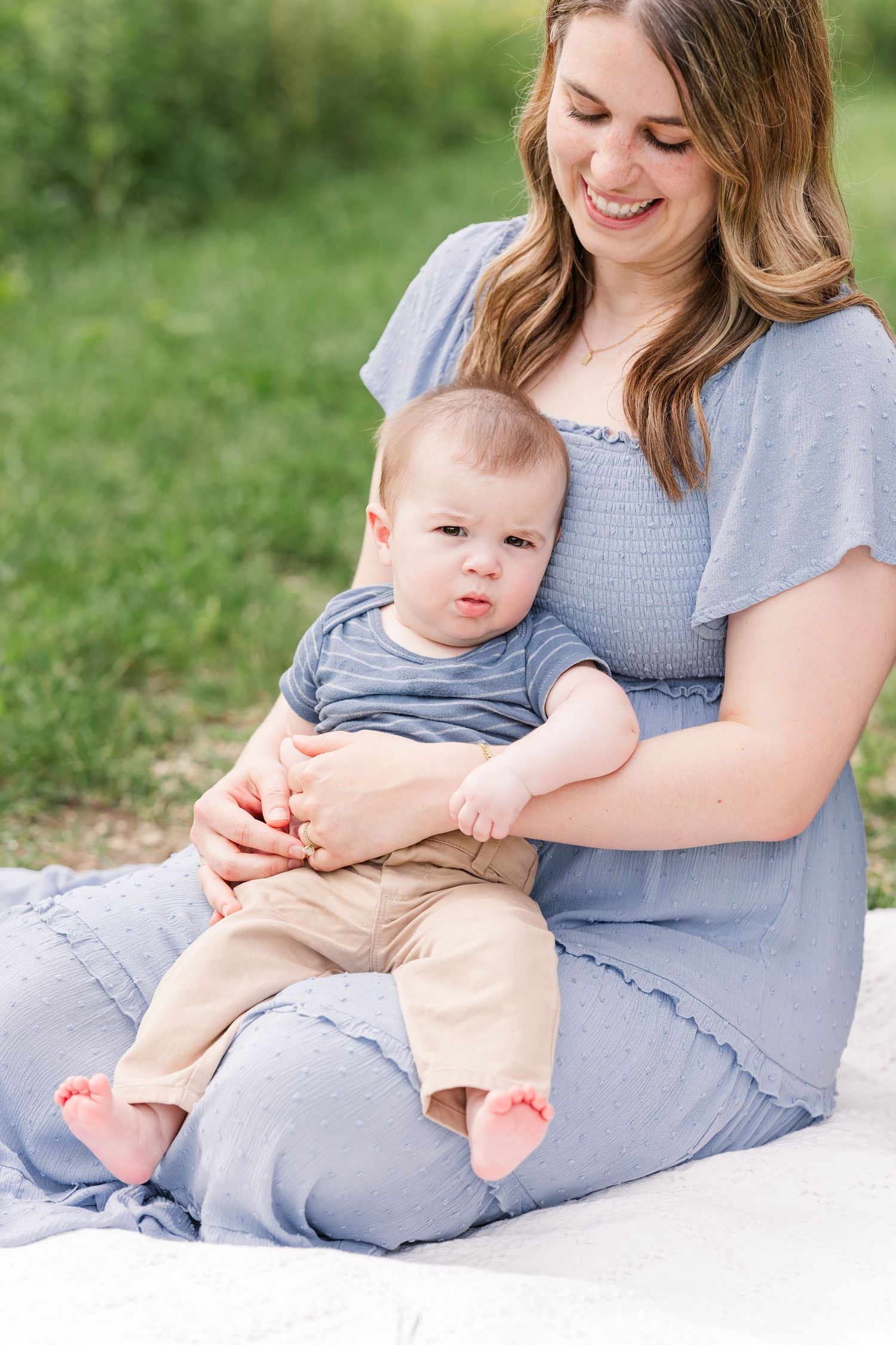 Smiling mother sitting outdoors on white blanket holding baby boy in blue onesie and tan pants in green park setting.