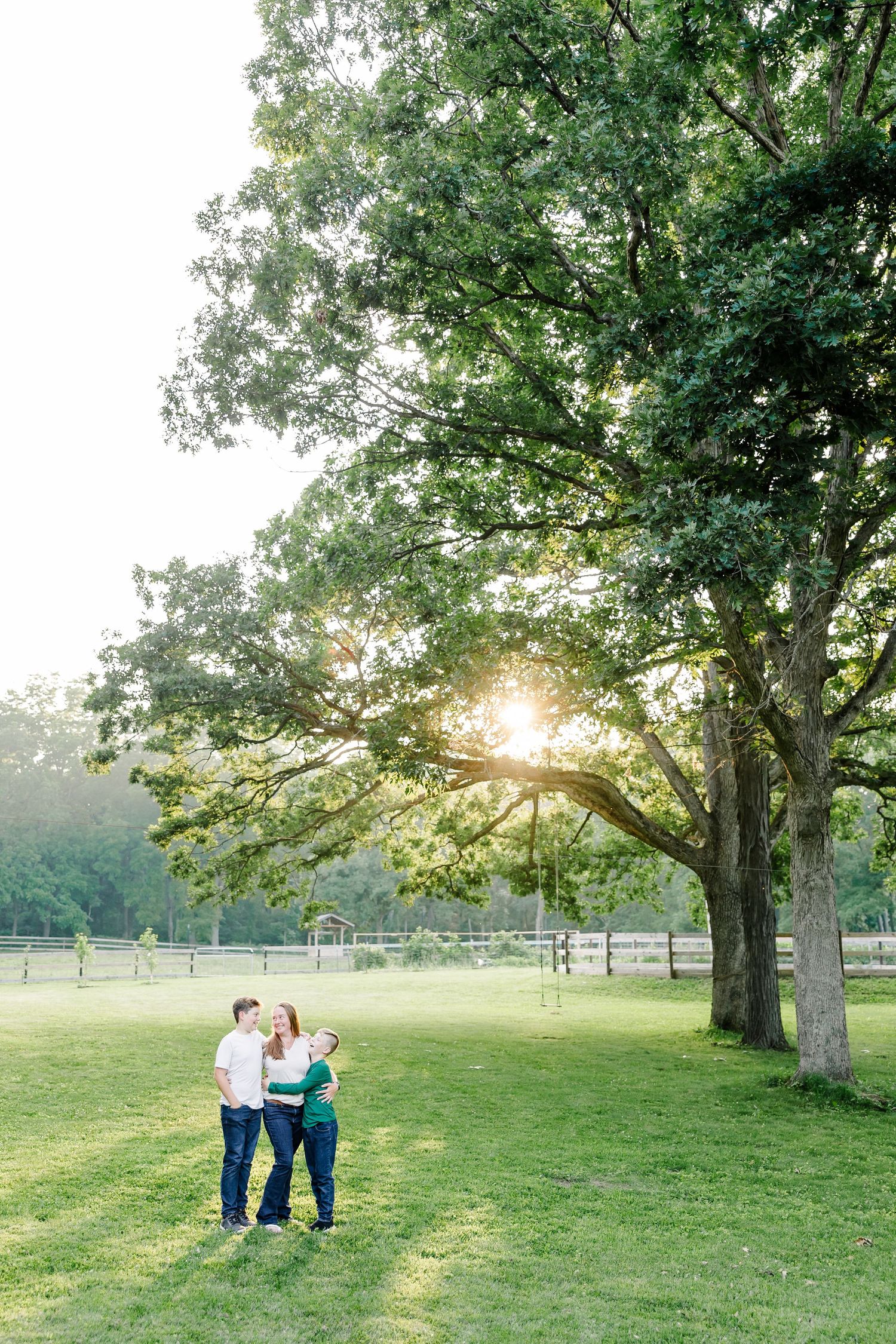 Family of three embracing on green lawn beneath large oak tree with golden sunset light behind them.