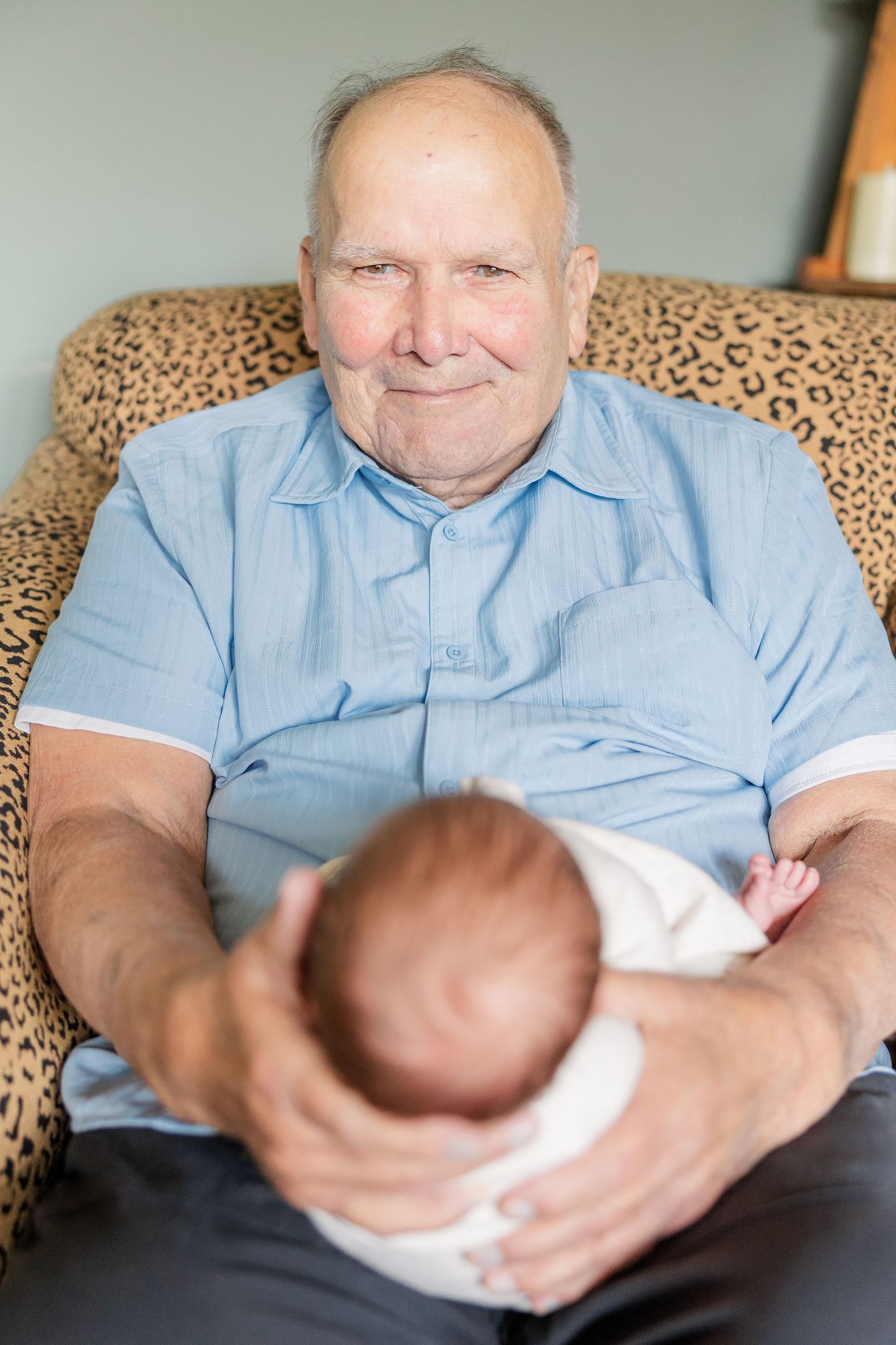 An elderly grandfather smiles warmly while cradling his newborn grandchild in a leopard print chair.