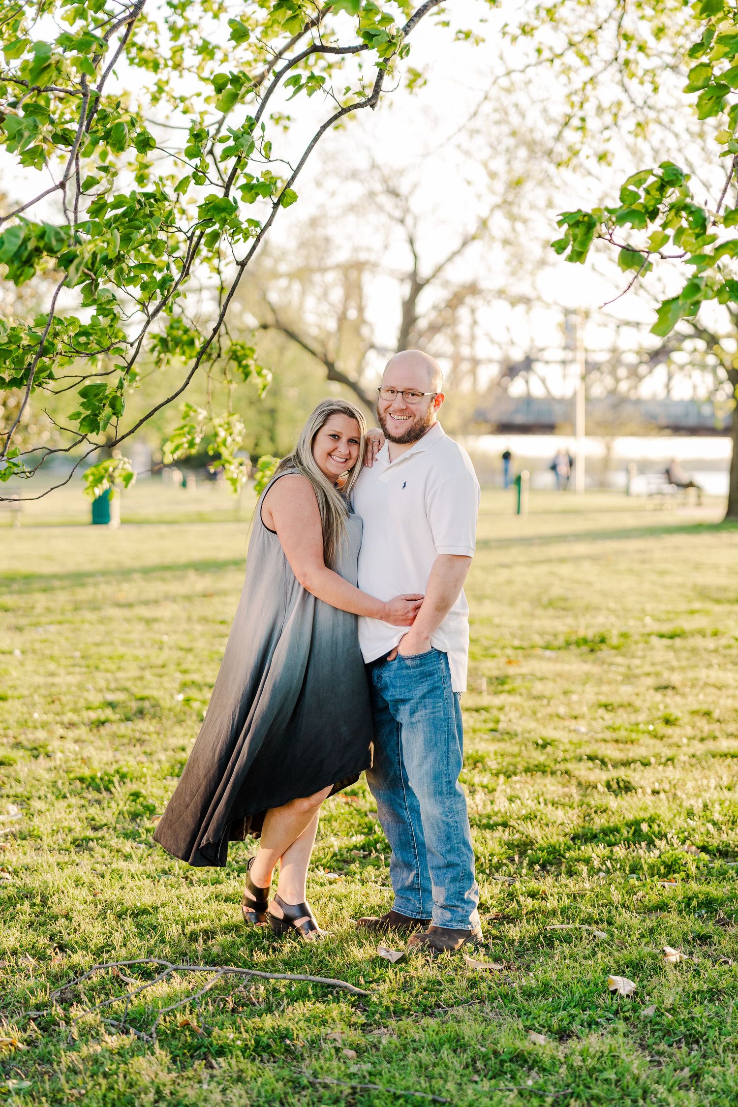 A couple poses together on a grassy lawn with spring trees and a bridge in the background.