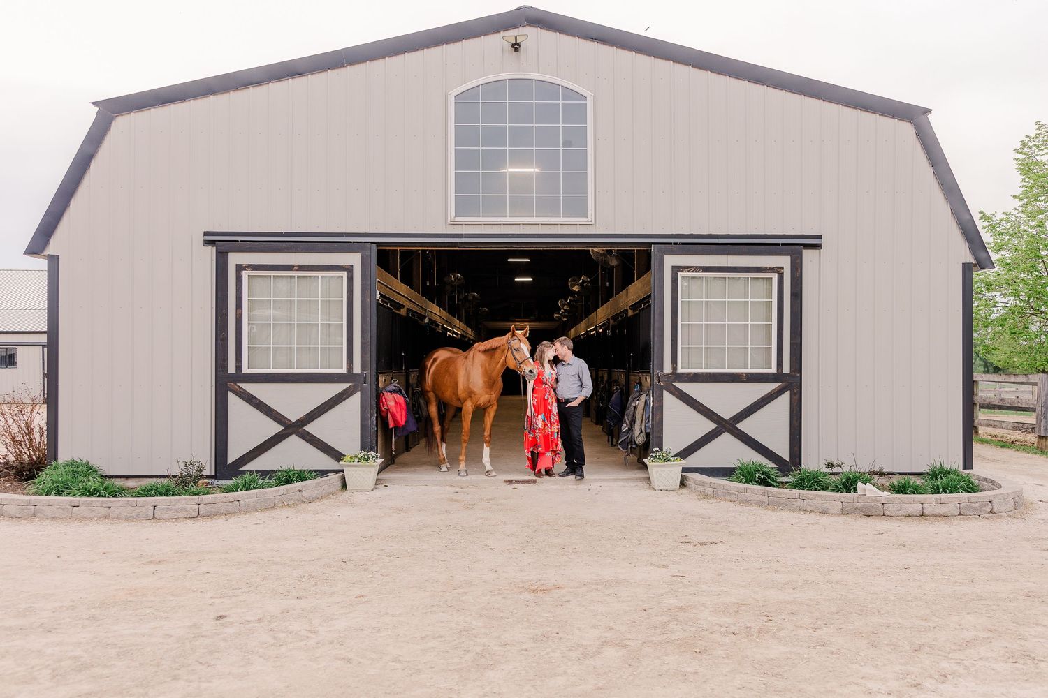 Large beige horse barn with black trim and windows, showing horses inside the doorway.