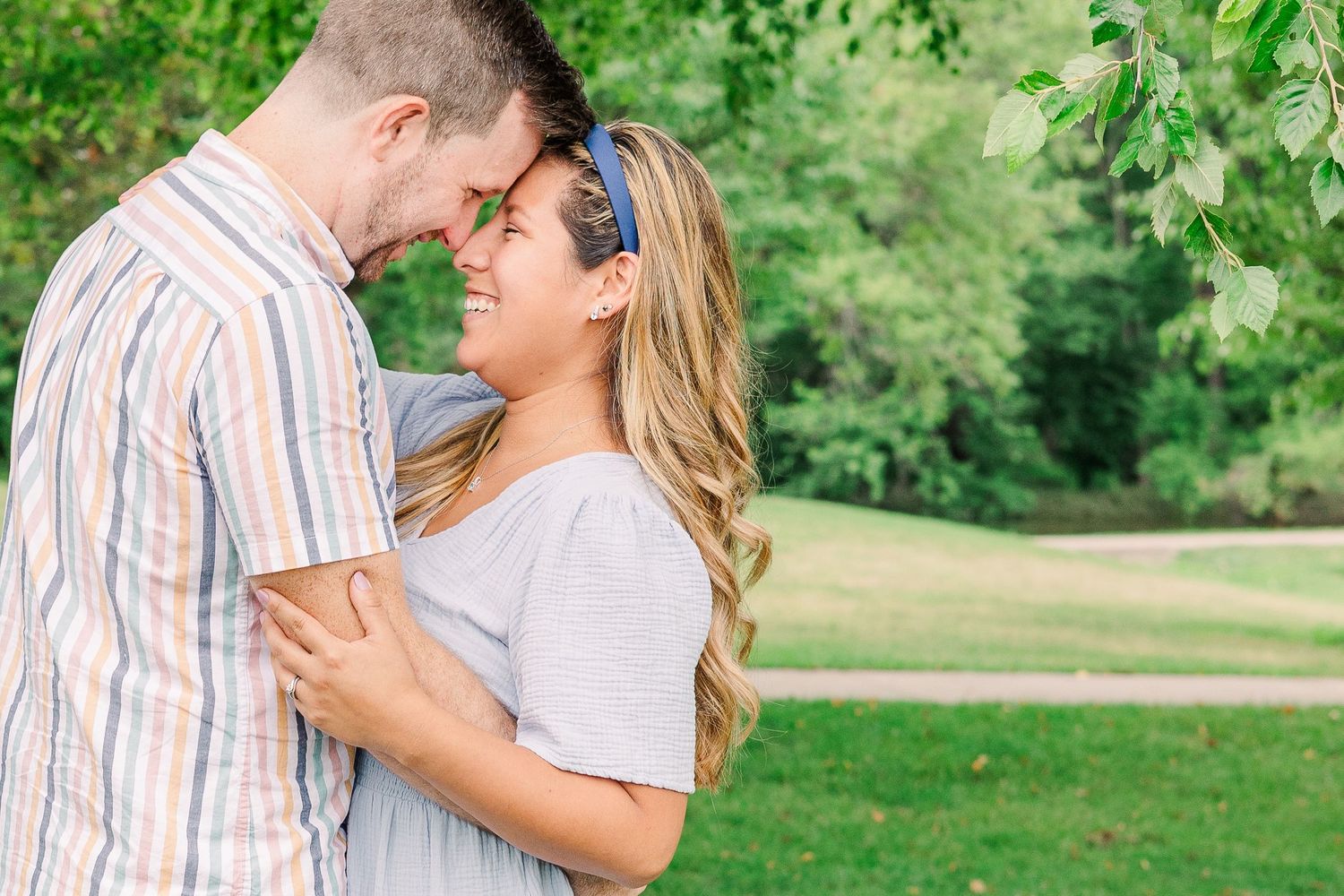 A tender embrace between a couple standing in a sunlit park with green foliage in the background.