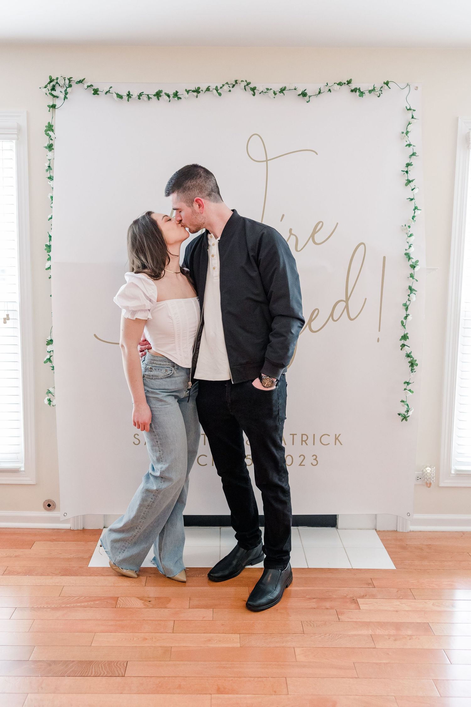 Engaged couple posing in front of elegant white backdrop with green vine border.