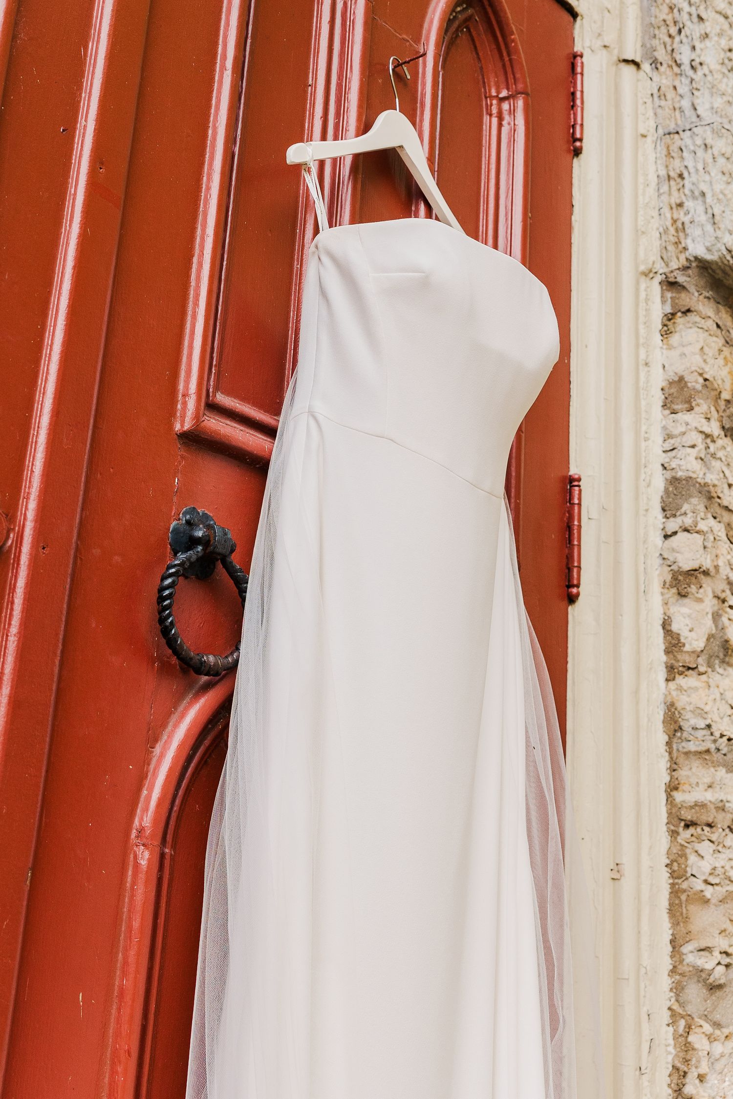 Close-up detail of a white wedding gown hanging on a red church door with an iron ring handle.
