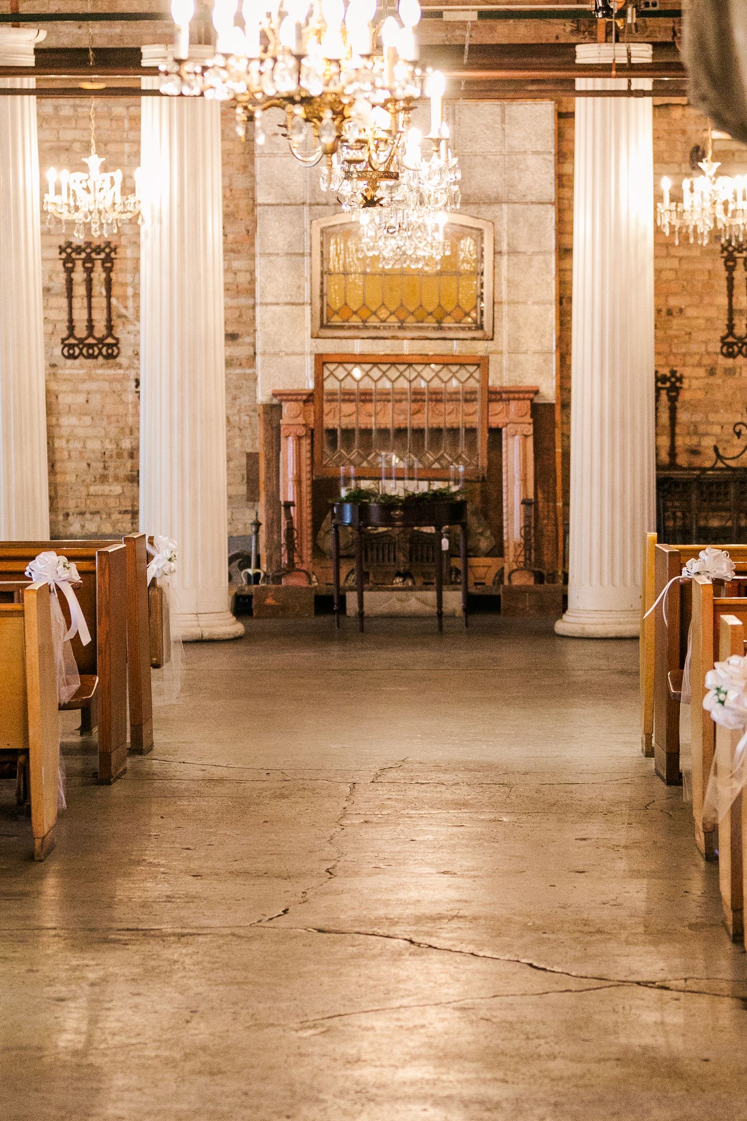 Empty ceremony aisle with wooden pews and crystal chandeliers in rustic brick venue.