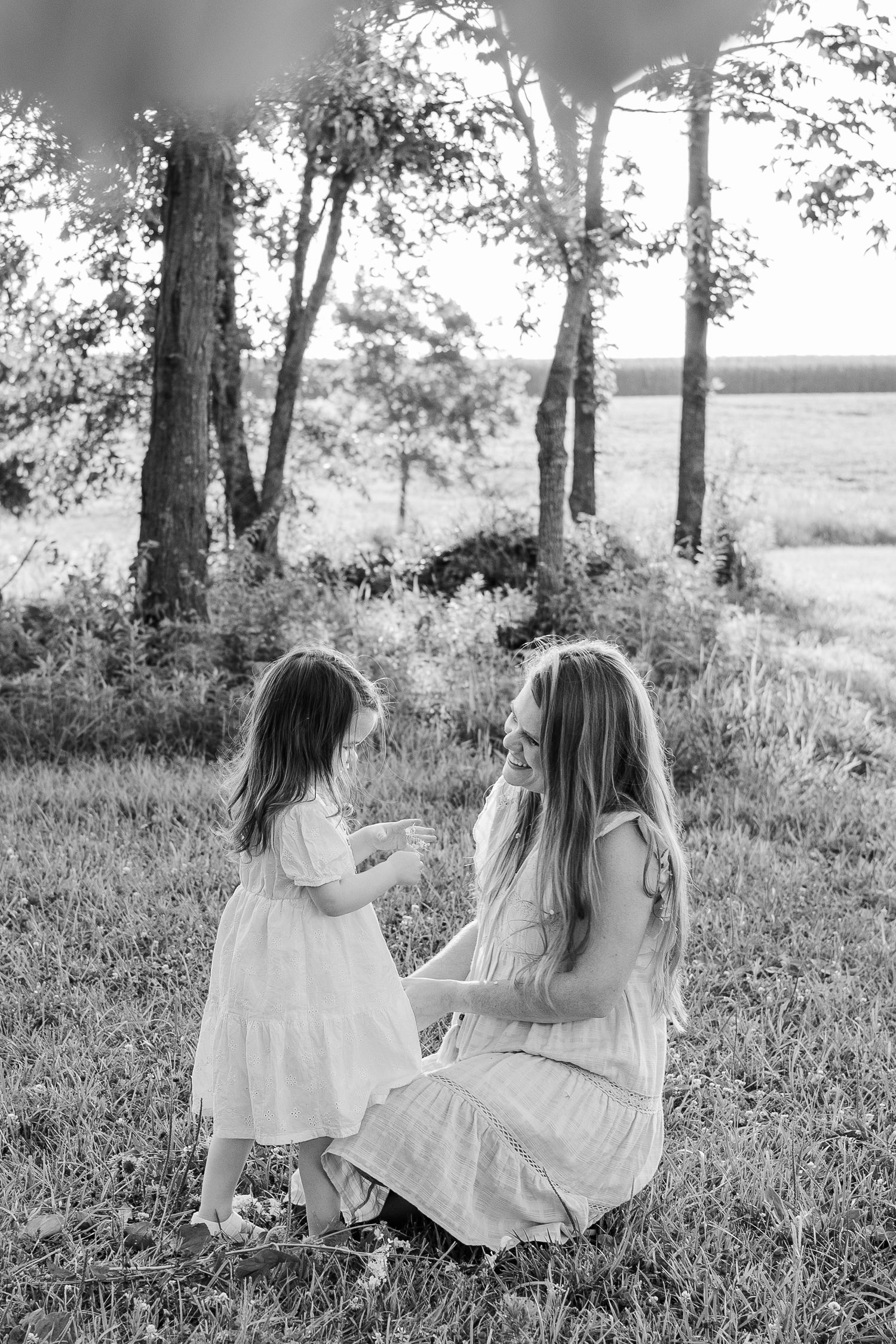 Black and white image of two people in white dresses sitting together on grass near trees.