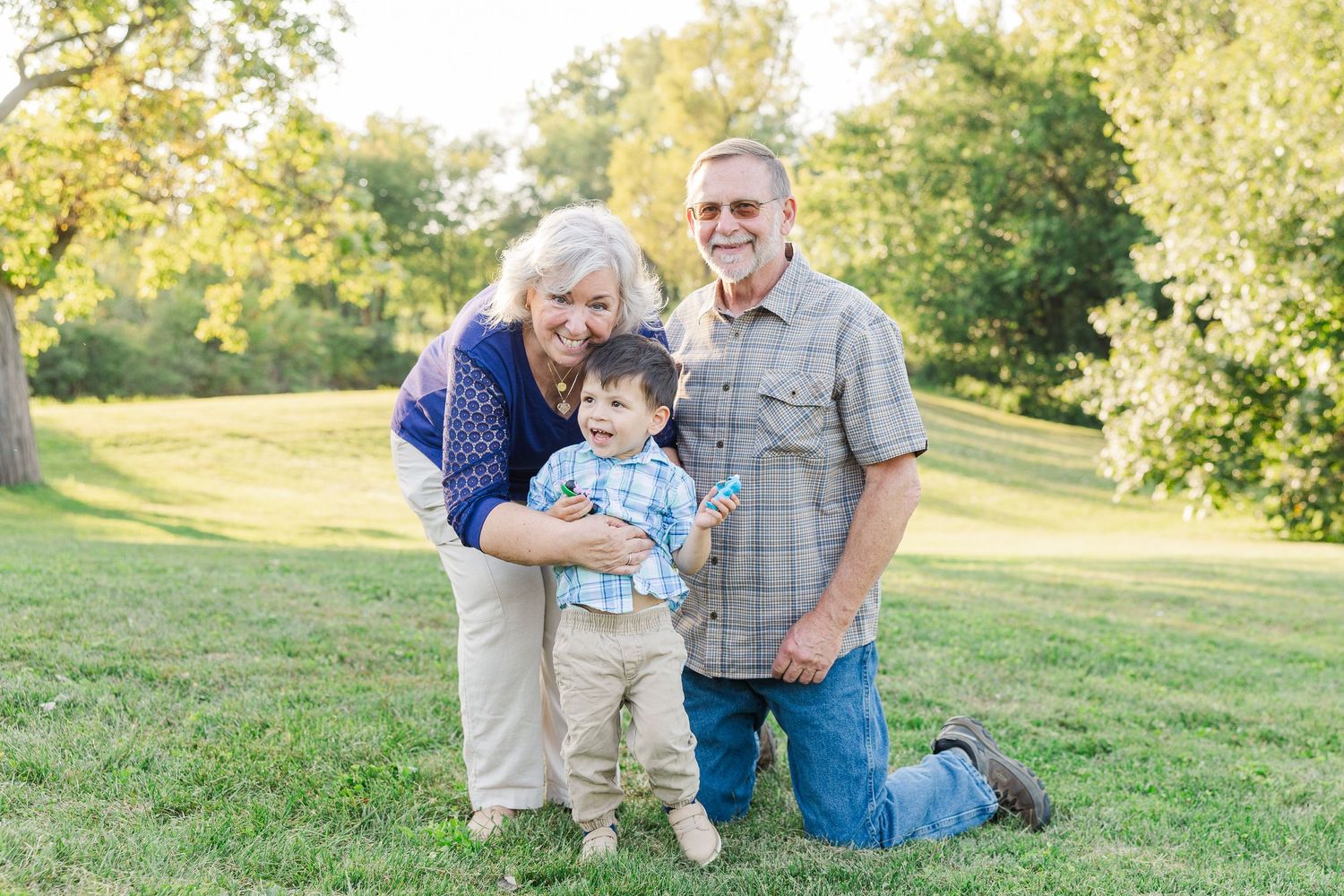 Three generations pose together outdoors on a sunny day in a grassy park setting.Sorg family portraits at Shamrock Hills Park in Crystal Lak