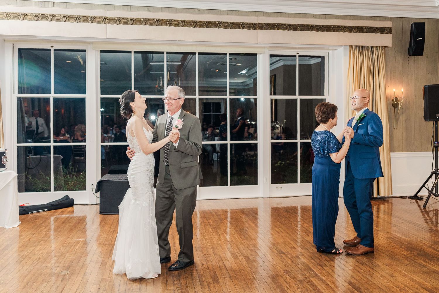Couples dancing on wooden floor at elegant reception venue with large windows.